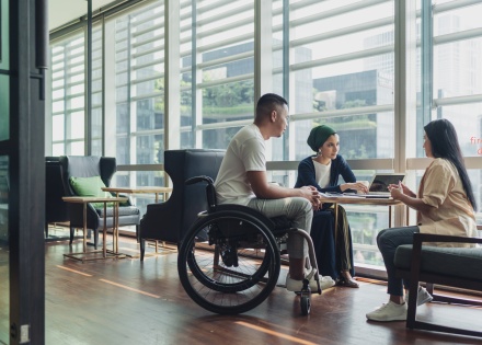 Employee in wheelchair works alongside his to co-workers at table by window.