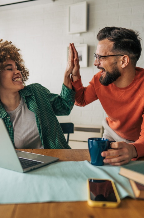 Man holding coffee high-fives his female colleague as they sit at a table in front of laptop.